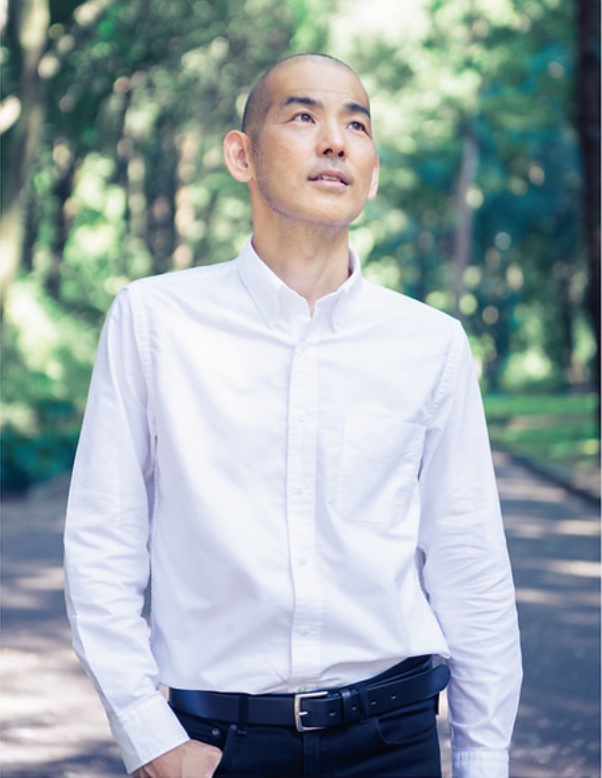 木山裕策さん。白いシャツで木立の道に立ち、空を見上げている